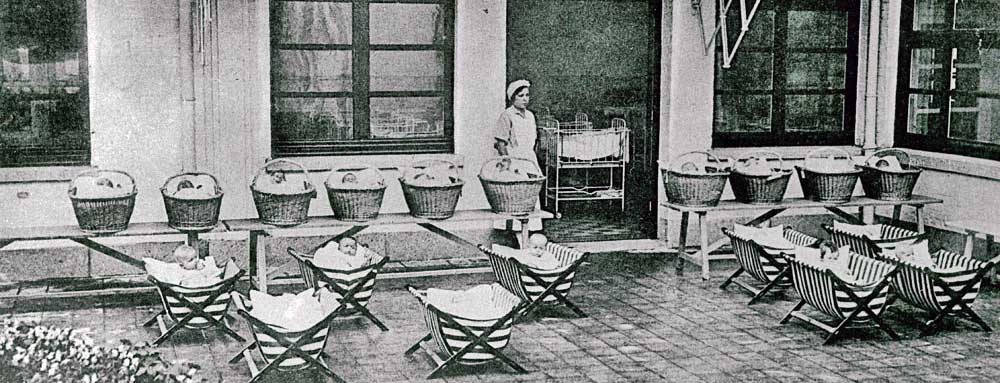 <p>Une nurse pose dans la cour, entourée de 10 paniers à bébés et 9 berceaux.</p>